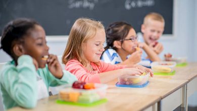 مجموعة متعددة الأعراق من الأطفال في سن الابتدائية يجلسون على مكاتبهم ويأكلون وجبات غداءهم الصحية.